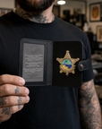 A tattooed man holds open a black leather badge holder displaying a gold sheriff-style badge with a blue and yellow palm tree emblem and a gray insert card with engraved text, set against a blurred indoor background.