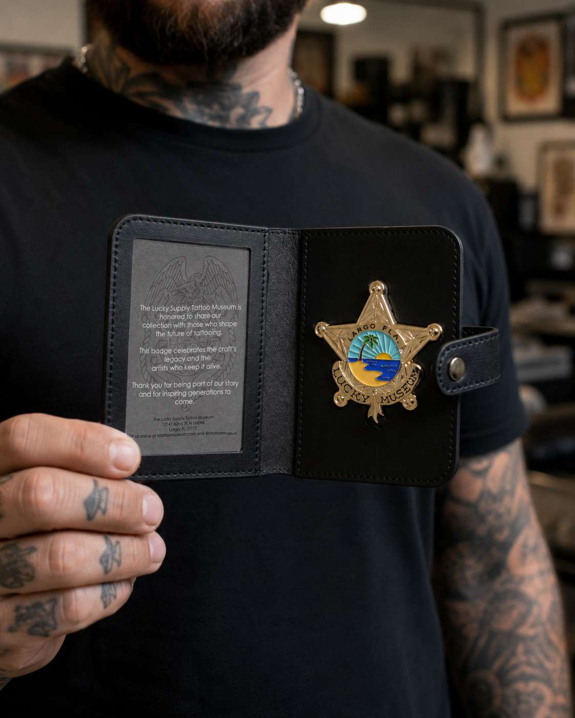 A tattooed man holds open a black leather badge holder displaying a gold sheriff-style badge with a blue and yellow palm tree emblem and a gray insert card with engraved text, set against a blurred indoor background.